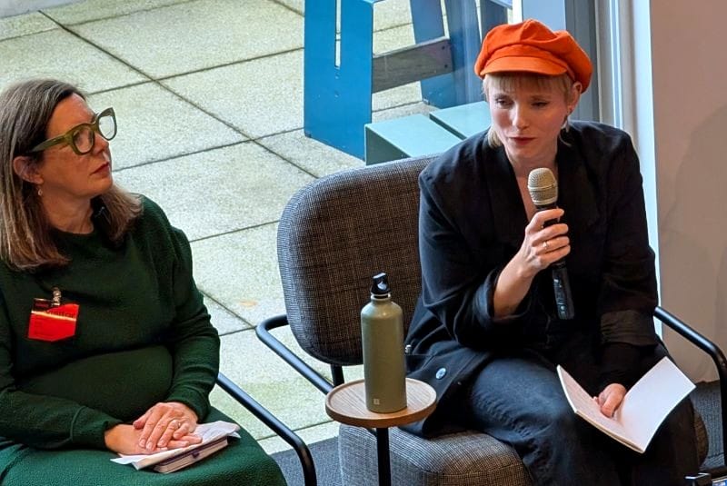 Tansy speaking into a microphone, wearing a black jacket and orange hat. Next to her sits Elisa in a dark green dress.